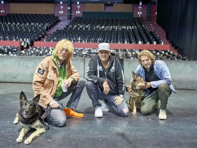 Drei Männer mit zwei Hunden hocken vor einer Tribüne