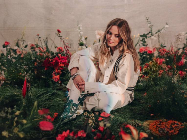 Blonde Frau sitzt zwischen roten Blumen.