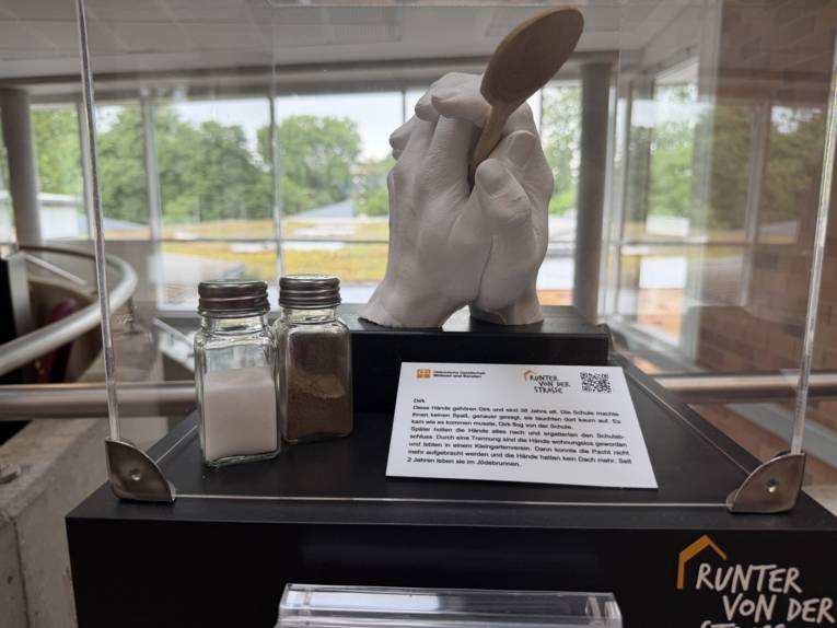 Zu sehen ist ein Foto einer Installation: Zwei Gipshände auf einem Sockel stehend halten einen Holzlöffel, daneben stehen ein Salz- und Pfefferstreuer.