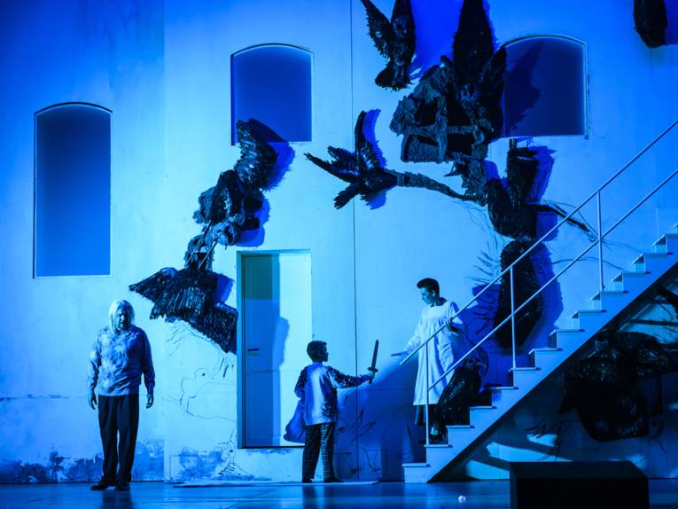 Auf einer Bühne ist vor einer Häuserfassade eine Treppe zu sehen. Links davon ist ein Mann Zeuge, wie ein Kind einer Frau, die auf der Treppe steht, einen Dolch überreicht.