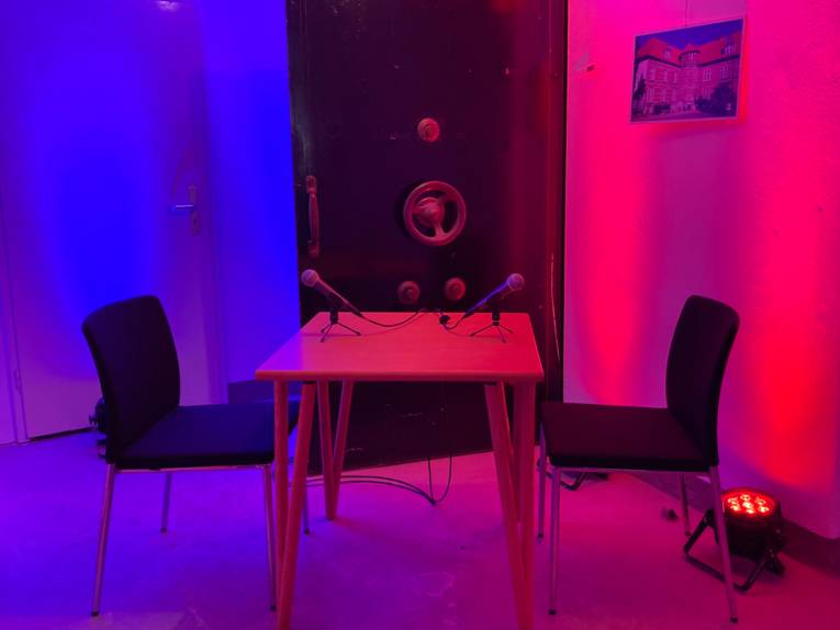 Neben der schweren Panzertür eines Tresors steht ein Tisch, daran zwei Stühle und darauf zwei Mikrofone. Scheinwerfer tauchen die Szene in Rot- und Blautöne.