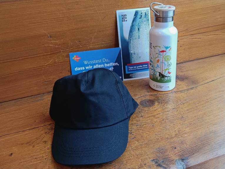 Eine Schirmmütze, eine Trinkflasche aus Edelstahl ein Flyer und eine Postkarte zusammengestellt auf einer Holzunterlage