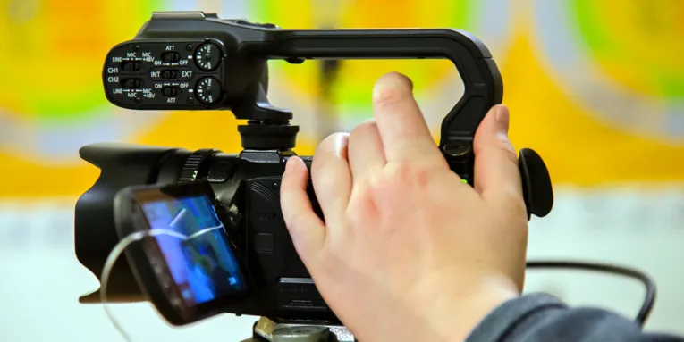 Eine Filmkamera filmt auf einem Stativ, die Hand einer Person ist an der Kamera.
