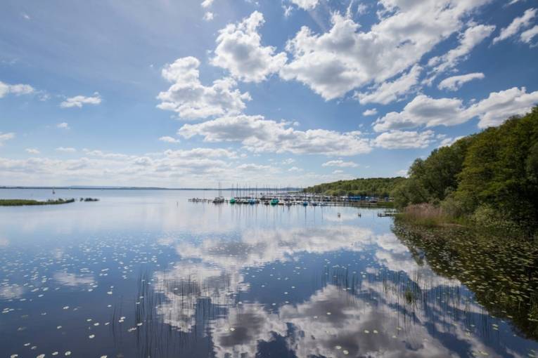 Blick über einen See, in dem sich Wolken spiegeln, im Hintergrund ein Steg mit Booten