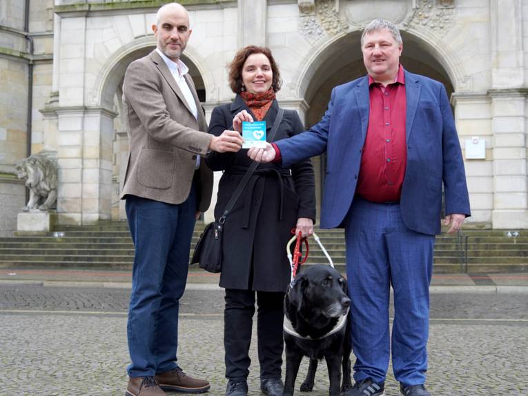 Zwei M&auml;nner und eine Frau in der Mitte halten einen blauen Aufkleber. Neben der Frau steht ein schwarzer Hund. Im Hintergrund sind Stufen und ein Eingangsportal mit zwei B&ouml;gen.