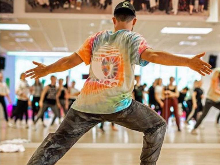 Ein Tänzer in buntem Shirt bringt anderen Tänzern in einem Tanzsaal eine Performance bei.