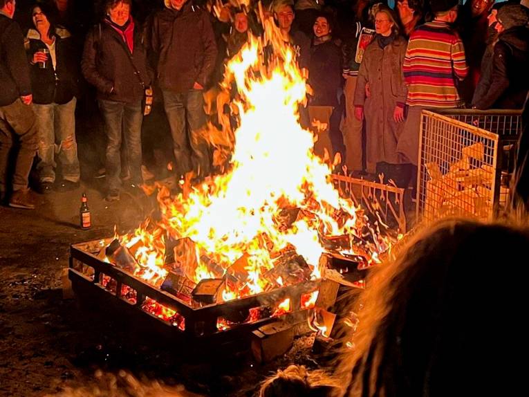 Menschen stehen um eine Feuerschale, in der es ordentlich lodert.