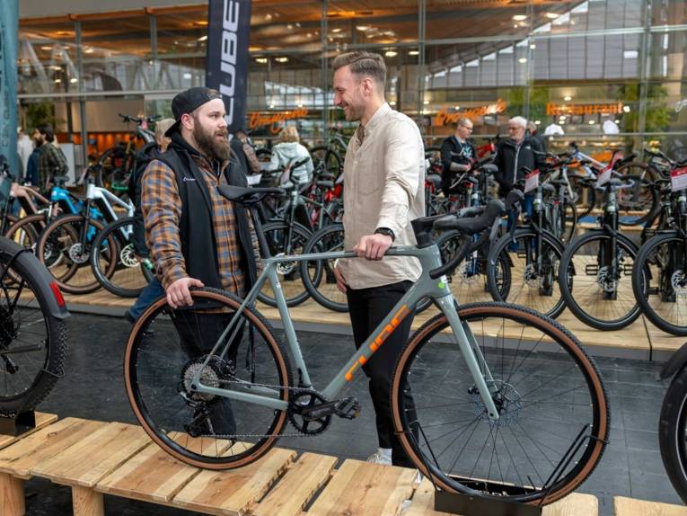 Zwei Männer mit einem Fahrrad in einer Messehalle, im Hintergrund weitere Fahrräder