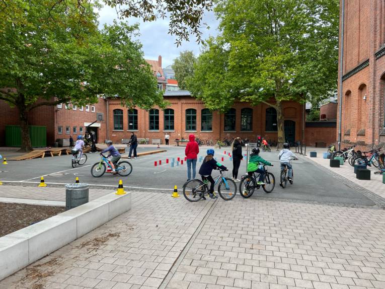 Auf dem Hof eines Backsteingebäudes fahren Kinder auf Fahrrädern entlang an Hindernissen, neben denen Erwachsene stehen.