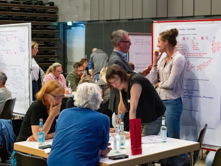 Menschen an Tischen im Gespr&auml;ch, einige sitzen, andere stehen, weitere stehen an Stellw&auml;nden mit beschriebenen Papieren.