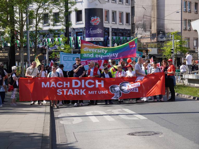 Auf einer Straße laufen Menschen und halten ein großes rotes Transparent vor sich und halten weitere hoch. Mach Dich stark steht auf dem großen Transparent.