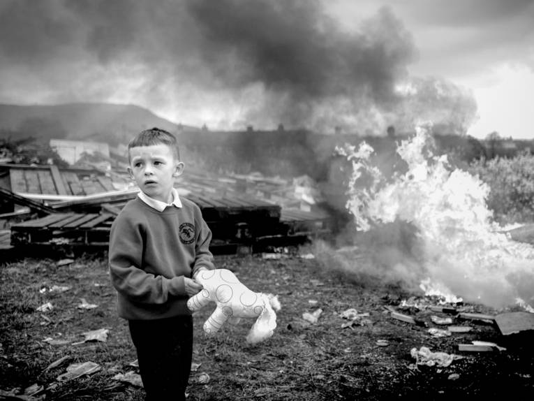 Zu sehen ist eine Fotografie von Toby Binder eines Schuljungen der ein Kuscheltier in der Hand hält. Er wendet sich vom Hintergrund weg, auf dem gerade ein Feuer ausbricht.