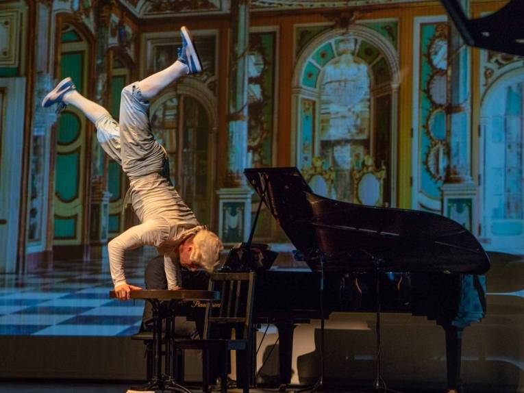 Zu sehen ist ein Breakdancer als Mozart verkleidet, wie er einen Handstand auf einem Klavier vollführt.