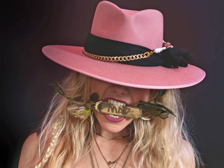 Blonde Frau mit pinkem Hut beißt auf ein Stück Gold, in das "Mad" eingraviert ist.