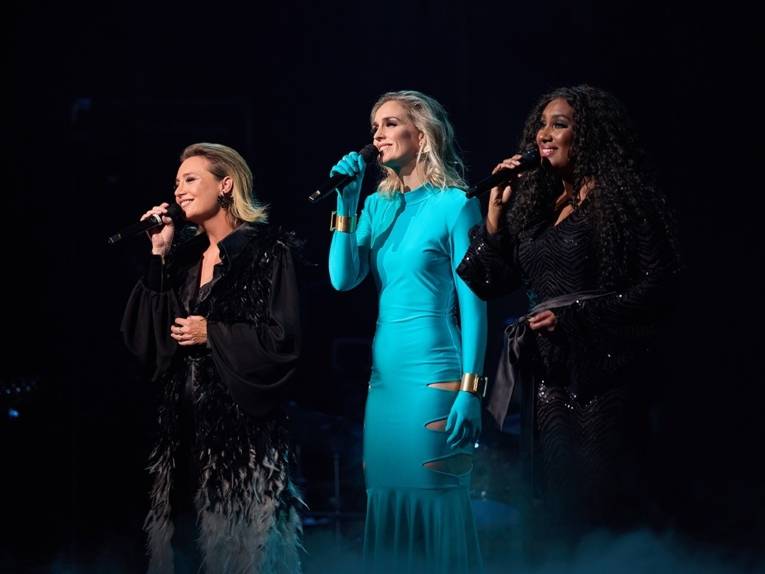 Zu sehen sind die drei Sängerinnen Ana Milva Gomes, Annemieke Van Dam und Wietske Van Tongeren in Abendkleidern auf der Bühne.