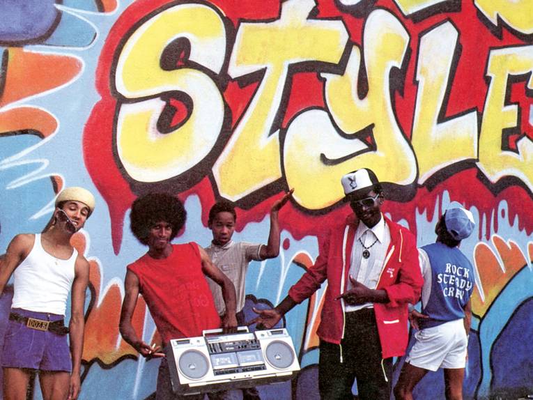 mehrere Personen in Partylaune mit einem großen Ghettoblaster stehen vor einer besprühten Wand, auf der das Wort Style zu sehen ist.