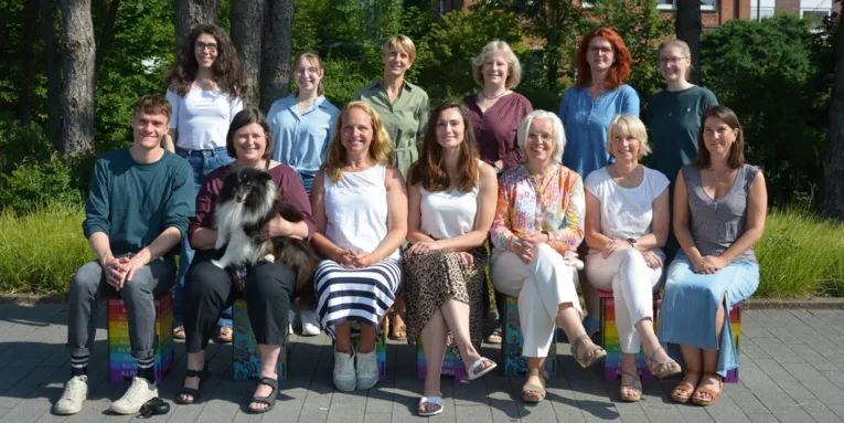 13 Personen und ein Hund sitzen oder stehen mit Blick in die Kamera für ein Gruppenfoto.