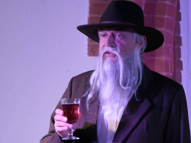 Mann mit weißem Bart, Hut und einem Glas Rotwein in der Hand.