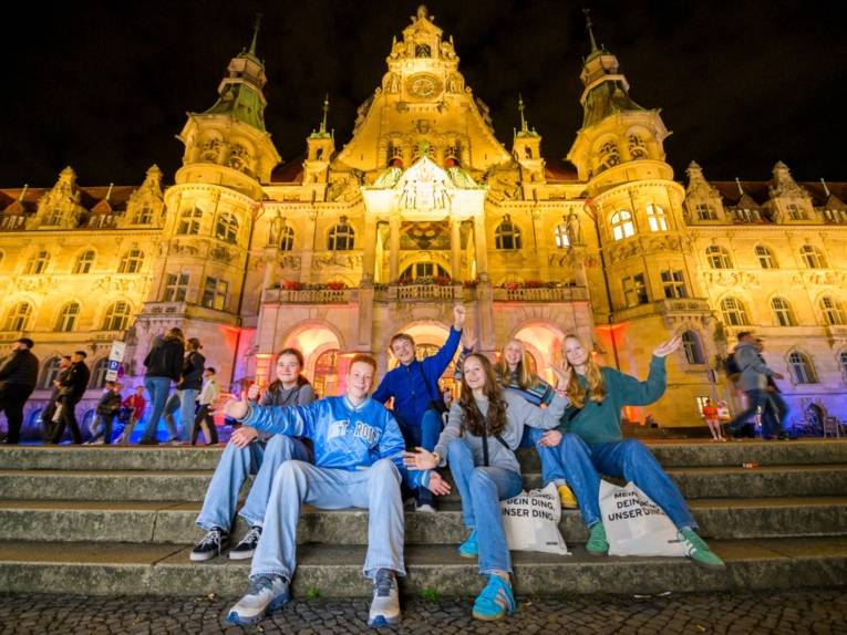 Eine Gruppe Jugendlicher sitzt auf der Treppe vor dem festlich beleuchtetem Neuen Rathaus in Hannover.