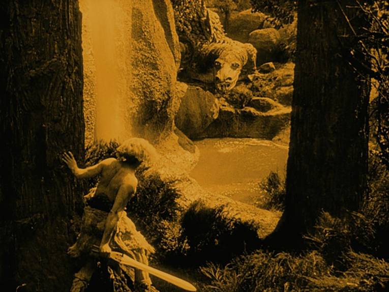 Eine Szene an einem Bergsee: ein Mann steht mit dem Rücken zur Kamera, er hält ein Schwert in der Hand. Auf der anderen Seite des kleinen Sees ein Drachen