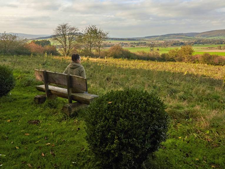 Eine Frau sitzt auf einer Bank und blickt auf die hügelige Deisterlandschaft im Herbst.