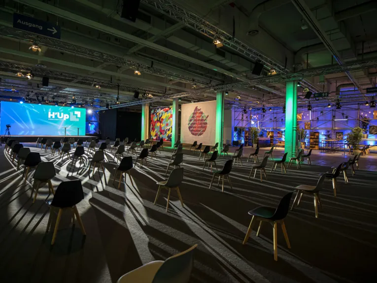 Beleuchtete, hybride Eventlocation der Deutschen Messe AG mit verstreuten Stühlen, Bühne mit „H’Up“-Logo und moderner Dekoration.