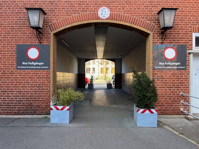 Ein Torbogen mit einem Durchgang in einem roten Klinkergebäude. Rechts und links des Bogens stehen Pflanztröge, darüber sind am Haus Schilder "Nur Fußgänger" angebracht. Hinter dem Durchgang ist Fußweg und der Ausschnitt eines weiteren Gebäudes zu sehen.