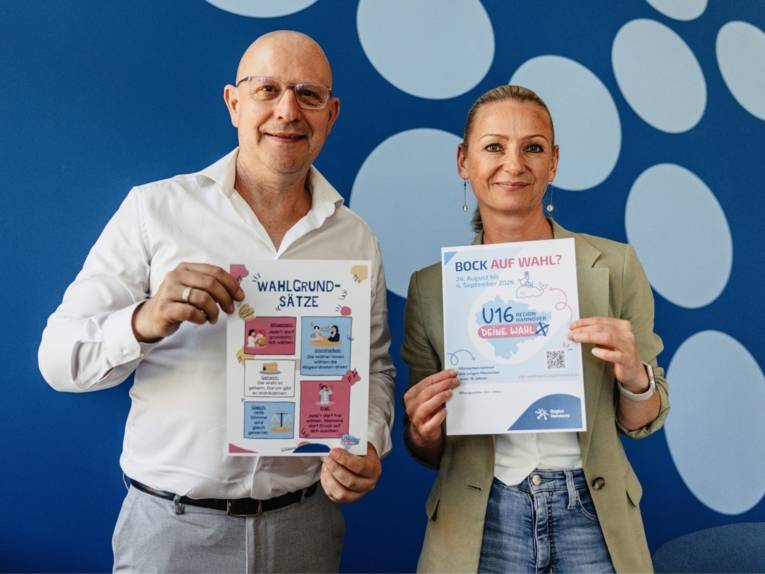 Ein Mann mit Glatze und eine blondhaarige Frau stehen nebeneinander und halten Zettel in der Hand mit Informationen zu der U16-Wahl.  Beide lächeln in die Kamera.