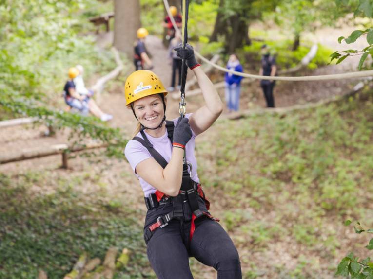 Eine Person hängt an einem Seil, im Hintergrund sind weiterer Personen und Klettermöglichkeiten
