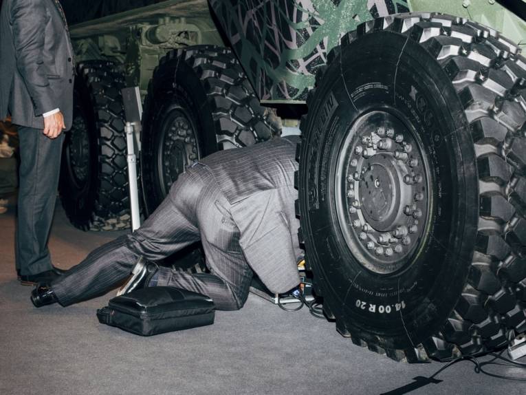 Zu sehen ist eine Fotografie von Nikita Teryoshin: Ein Mann im Anzug kriecht zwischen großen Reifen eines militärischen Fahrzeuges. Ein anderer Mann