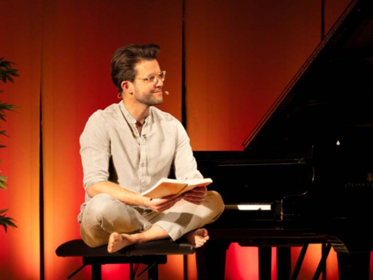 Dennis Alexander Volk sitzt an einem Klavier und liest aus einem Buch vor.