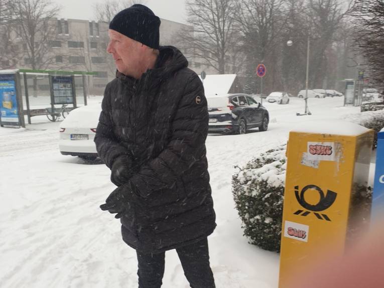 Ein Mann in Winterjacke und Mütze auf einer verschneiten Straße.