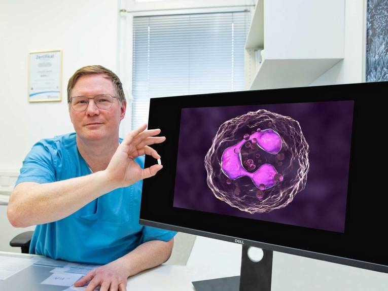 Ein Mann im blauen Kittel sitzt neben einem Bildschirm mit der Abbildung einer Immunzelle und hält eine weiße Tablette in der Hand.