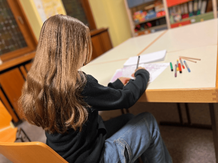 Ein Kind mit langen blonden Haaren sitzt an einem Tisch und schreibt etwas auf. Auf dem Schreibtisch sind mehrere vollgeschriebene Blätter Papier und verschiedenfarbige Stifte.