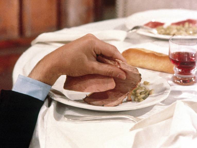 Eine Hand greift von unten auf einen Tellen und nimmt eine Scheibe Fleisch