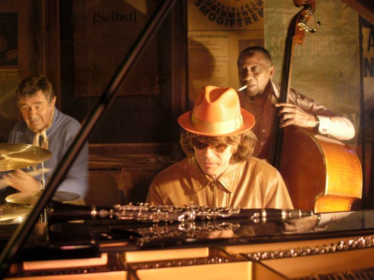 Warme Szene in einem kleinen Jazzclub: Im Vordergrund sitzt eine Person mit Hut und Sonnenbrille an einem Klavier. Dahinter spielen zwei weitere Personen Schlagzeug und Kontrabass, eine mit einer Zigarette im Mund. Das Licht ist gedämpft und bernsteinfarben, im Hintergrund hängen Plakate an den Wänden.