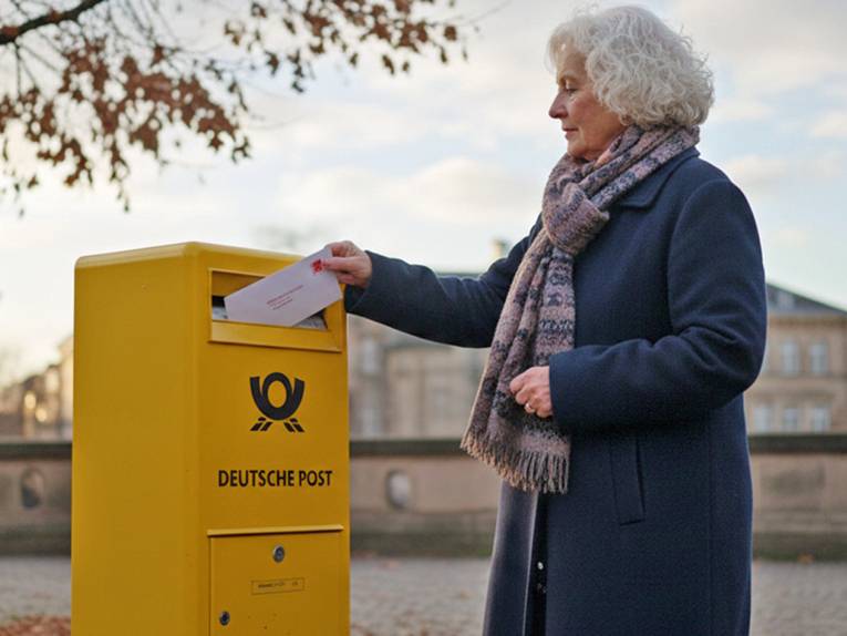 Ältere Frau wirft einen Brief in einen Briefkasten.