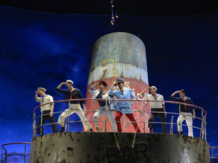 Sechs Schauspielende in Matrosen-Uniform auf einer Bühne, die einen Schornstein eine Kreuzfahrtschiffs darstellt.