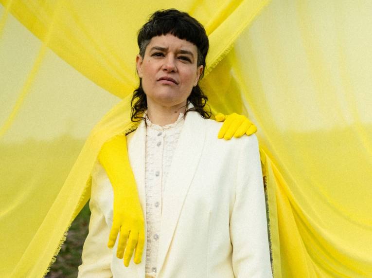 Frau mit zurückgestecktem Haar in Blues und Sakko, über ihren Schultern liegen zwei "Extra-Arme" mit Gummihandschuhen.