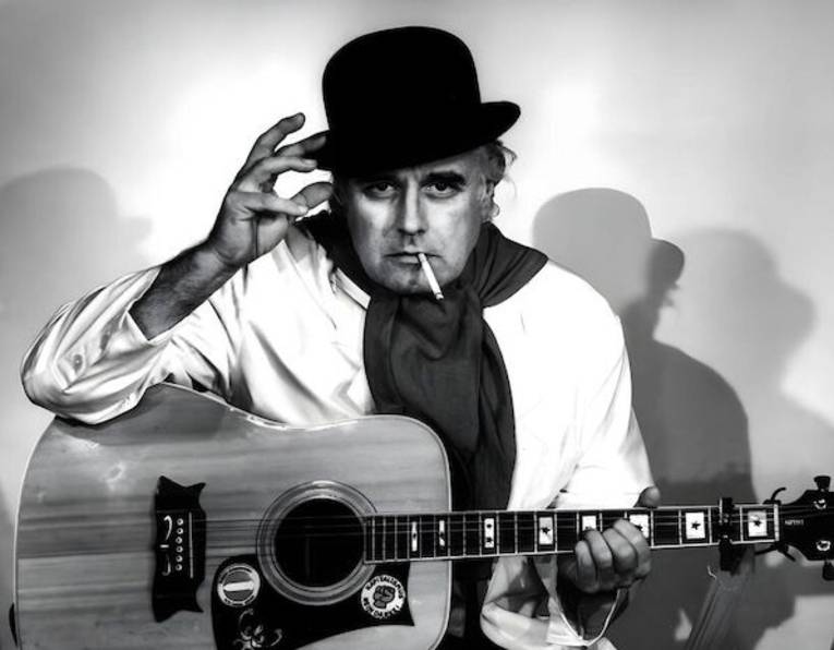 Mann mit Hut, Halstuch, Zigarette im Mund und Gitarre auf dem Schoß