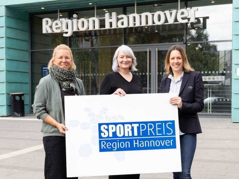 Drei Frauen stehen vor einem Gebäude, über dem Eingang steht "Region Hannover" und sie halten ein Schild in der Hand mit der Aufschrift "Sportpreis der Region Hannover".