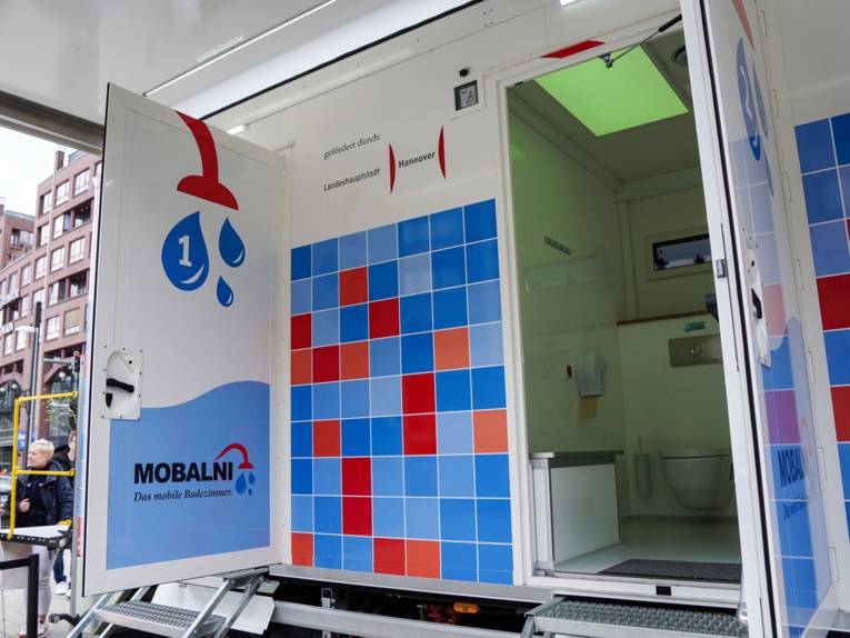 Ein blau-rot gekachelter Anhänger mit zwei geöffneten Türen, darin sind eine Ablage und eine Toilette zusehen.