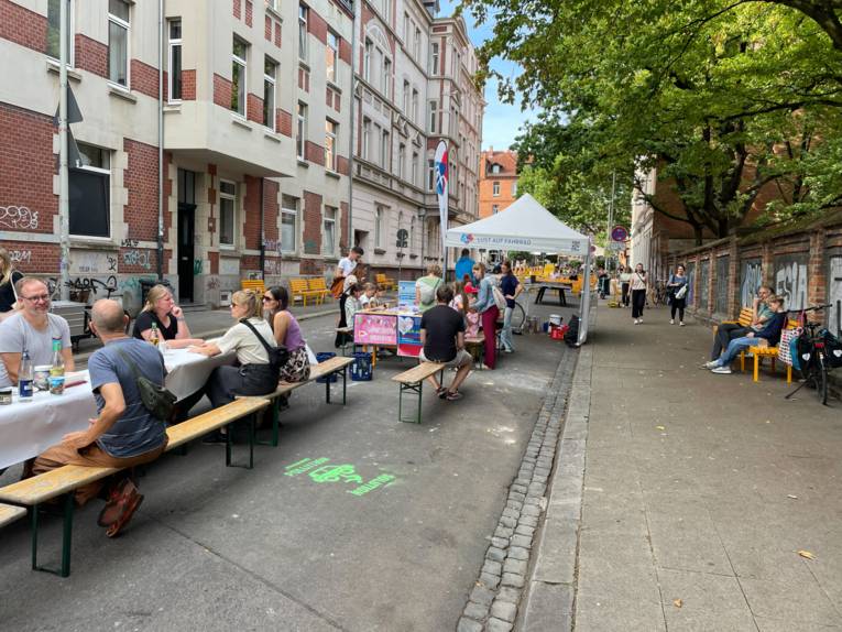 Auf einer Straße stehen Bänke und Tische, an denen Menschen sitzen und sich unterhalten.