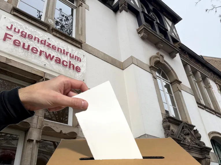 Eine Hand wirft einen Umschlag in eine Wahlurne. Im Hintergrund ein Jugendzentrum "Feuerwache".