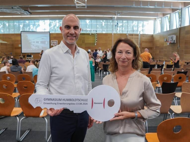 In einer Halle mit Stuhlreihen udn Menschen im Hintergrund stehen vorn ein Mann und eine Frau und halten einen überdimensionalen Schlüssel in den Händen.