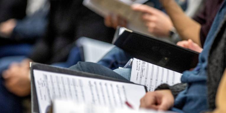 Menschen sitzen in Stuhlreihen nebeneinander und haben Mappen mit Noten auf dem Schoß.