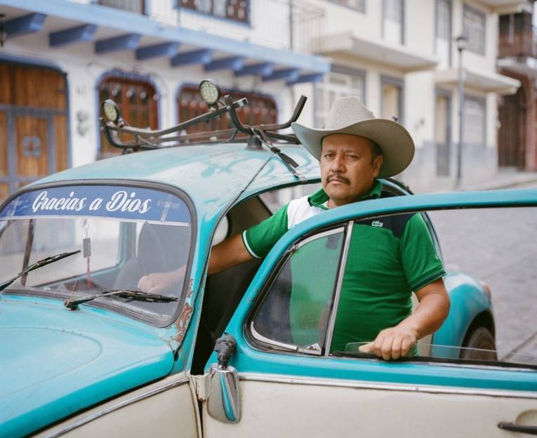 Ein Mexikaner mit Hut steht neben seinem VW-Käfer, dessen Fahrertür geöffnet ist