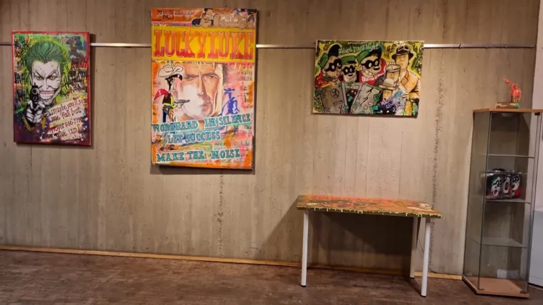 Drei knallig-bunte Bilder h&auml;ngen an der grauen Wand. Zu sehen sind unter anderem der Joker, Lucky Luke und die Panzerknacker. Vor der Wand steht ein bemalter Tisch und eine Vitrine. In der Vitrine steht ein Koffer.