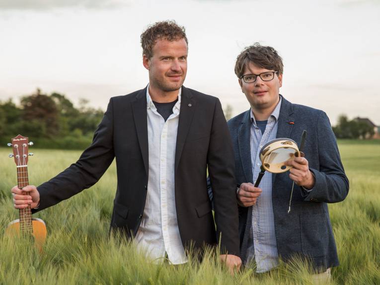 Zwei Männer mit Gitarre bzw. kleiner Trommel auf einer Wiese mit hüfthohem Gras