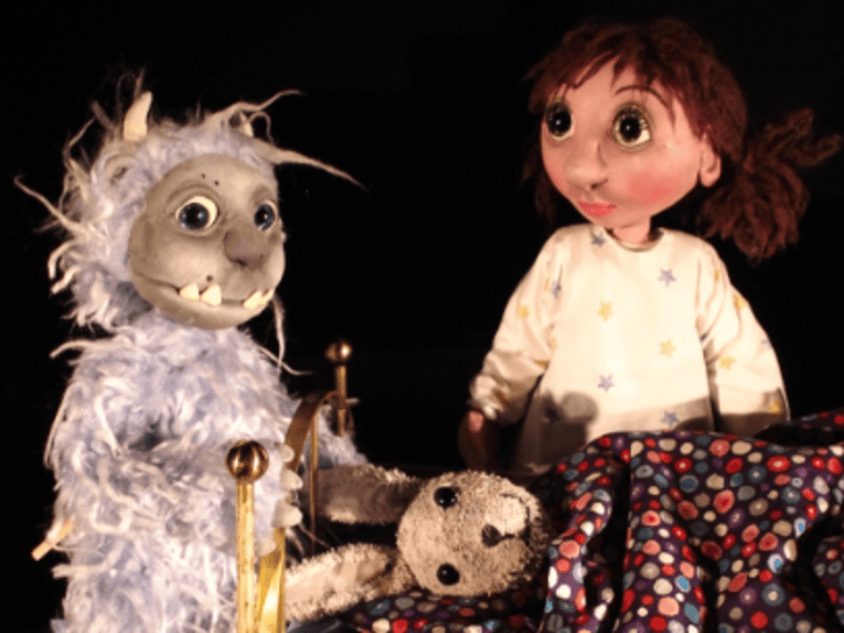Zu sehen ist ein Puppenspiel: Ein Monster und ein Mädchen stehen hinter einem Bett, auf dem unter einer Decke ein Hase liegt.